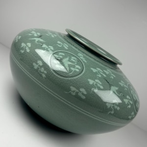 May include: A celadon ceramic vase with a crackle glaze. The vase features a design of birds and floral patterns in white. The vase has a rounded body and a flat, circular opening at the top. The overall shape is oval.