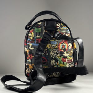 Puede incluir: Una mochila peque&ntilde;a con un estampado colorido de ilustraciones vintage de Mickey Mouse, que incluye el texto "Mickey Mouse wrist watch $2.95". La bolsa tiene correas, un asa superior y bordes negros, mostrada desde la parte posterior.