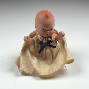May include: A small doll with a pale, molded head, blue painted eyes, and a tiny red mouth. The doll wears a beige, textured fabric garment that is gathered at the neckline and features a small, dark blue ribbon bow centered at the front.