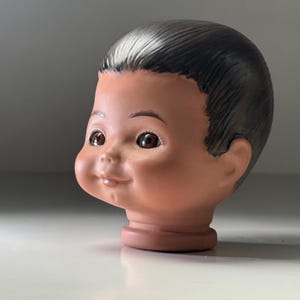 May include: A vintage doll's head with a smiling expression. The head is made of porcelain or a similar material, with brown eyes, rosy cheeks, and dark hair. The head is a light peach colour.
