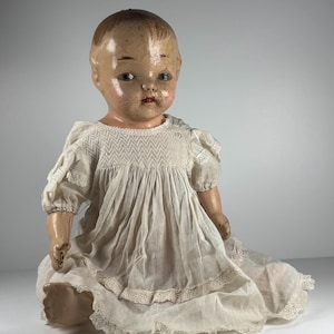 May include: A vintage composition doll with a painted face, brown hair, and dark eyes. The doll wears a white, short-sleeved dress featuring intricate smocking across the chest, a gathered skirt, and delicate lace trim along the hem and sleeves.