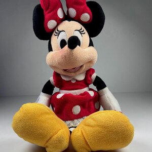 May include: A plush Minnie Mouse doll, featuring a red dress with white polka dots, a matching bow, and yellow shoes. The doll has black ears and a smiling face.