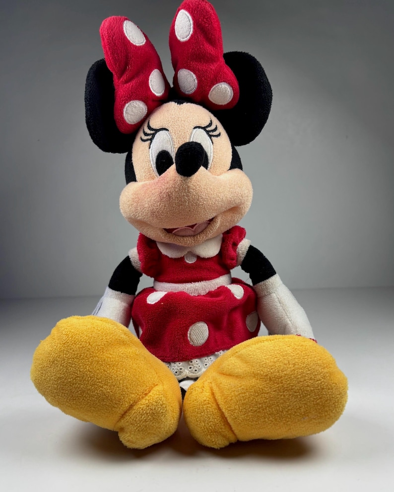 May include: A plush Minnie Mouse doll, featuring a red dress with white polka dots, a matching bow, and yellow shoes. The doll has black ears and a smiling face.