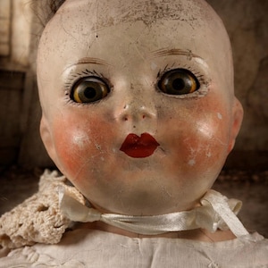 May include: Close-up of an antique doll with a cracked porcelain face. The doll has large, expressive eyes, rosy cheeks, and red lips. It is wearing a white dress with a lace collar and a satin ribbon.