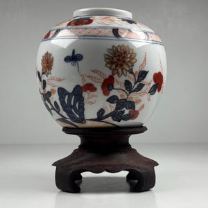May include: A white ceramic vase with floral designs in orange, red, and blue, set on a dark wooden stand. The vase features a rounded shape with a narrow neck and intricate patterns.