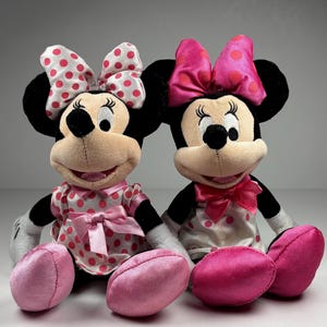 Op de afbeelding: Twee pluche Minnie Mouse poppen. De ene pop draagt een witte jurk met roze stippen en een bijpassende strik. De andere pop draagt een witte jurk met roze stippen en een roze strik. Beide poppen hebben zwarte oren, zwarte ogen en roze voeten.