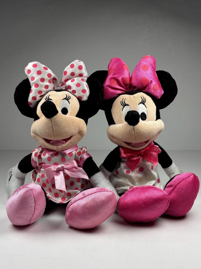 Op de afbeelding: Twee pluche Minnie Mouse poppen. De ene pop draagt een witte jurk met roze stippen en een bijpassende strik. De andere pop draagt een witte jurk met roze stippen en een roze strik. Beide poppen hebben zwarte oren, zwarte ogen en roze voeten.