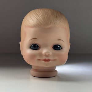 Pode incluir: Uma cabeça de boneca vintage com uma expressão serena. A cabeça é feita de um material pálido cor de pele, com olhos azuis, cílios delicados e um sorriso sutil. O cabelo é penteado de forma simples e suave.