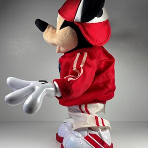 May include: A plush Mickey Mouse doll wearing a red and white baseball cap and a red jacket with white stripes. The doll has white gloves and white shoes with red stripes and soles. The doll is a classic Disney collectible.