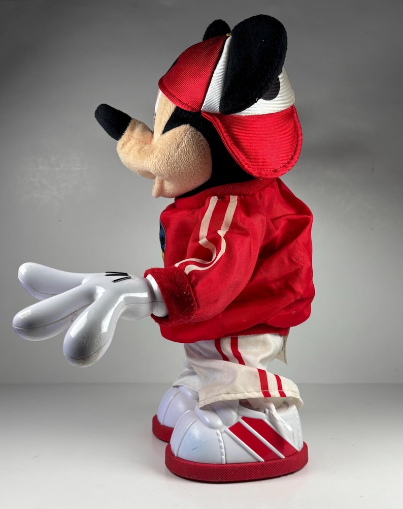 May include: A plush Mickey Mouse doll wearing a red and white baseball cap and a red jacket with white stripes. The doll has white gloves and white shoes with red stripes and soles. The doll is a classic Disney collectible.