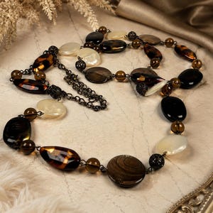 May include: A long necklace featuring a dark metal chain interspersed with various beads. The beads include smooth cream-colored discs, translucent brown glass spheres, and polished dark brown and black oval stones with marbled patterns.
