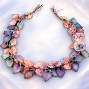 Può includere: Una collana caratterizzata da una serie di elementi in tessuto a forma di fiore nelle tonalità del rosa, viola e blu. I componenti floreali sono intervallati da piccole perline metalliche lungo un cordino, con estremità annodate in alto.