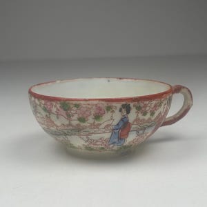 May include: A small, antique teacup with a delicate floral and landscape design. The cup has a red rim and handle, with a painted scene of a woman in traditional attire. The cup is likely made of porcelain.
