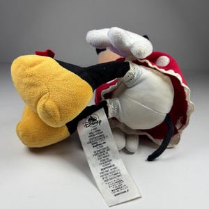 May include: A plush Minnie Mouse doll, lying on its side. The doll features a red and white polka-dot dress, white gloves, and yellow shoes. The tag reads "Disney".