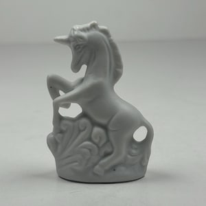 May include: A gray ceramic unicorn figurine. The unicorn is posed rearing up on its hind legs, with its front legs raised. The base features a decorative floral design. The figurine is a solid, matte gray color.