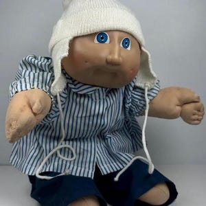 May include: A vintage doll with a tan face, blue eyes, and rosy cheeks. It wears a white knitted cap, a blue and white striped shirt, and navy blue trousers. The doll has soft, stuffed limbs.