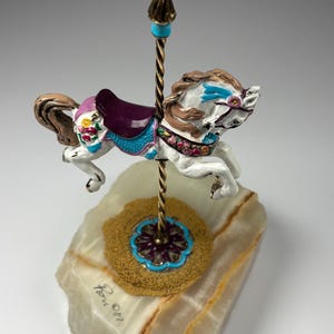 May include: A decorative carousel horse figurine. The white horse has brown and blue accents, a purple saddle, and floral details. It is mounted on a gold-coloured post with a turquoise accent, set on a marble base.