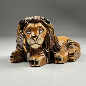 May include: A ceramic lion figurine in a resting pose. The lion features a brown body with gold and black accents on the mane, paws, and details. The figurine has a detailed, hand-painted appearance.