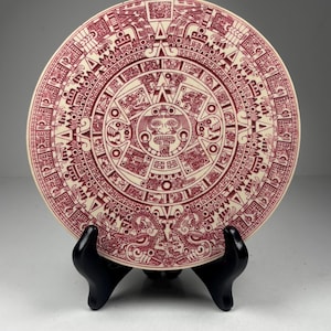 May include: A cream-colored decorative plate featuring a detailed Aztec calendar design in burgundy. The plate is round and displayed on a black stand, showcasing intricate patterns and symbols. A unique piece for home decor.