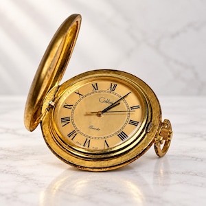 Può includere: Un orologio da tasca dorato con coperchio incernierato aperto, caratterizzato da un quadrante circolare con numeri romani neri, lancette nere e il testo Colibri e Quartz stampato sul quadrante. L'orologio include una piccola corona.