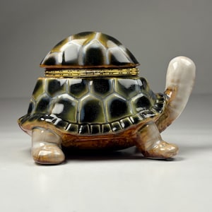 May include: A ceramic turtle figurine with a hinged lid. The turtle's shell is a mosaic of black, green, and brown, with a gold-colored band around the lid. The head and legs are a creamy beige color. The figurine is a decorative item.