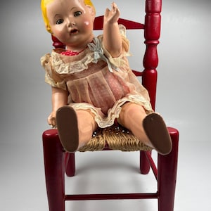 Puede incluir: Una muñeca vintage con cabello amarillo y mejillas rosadas, vestida con un vestido rosa con ribete de encaje, está sentada en una pequeña silla de madera roja con un asiento tejido. El brazo de la muñeca está levantado y tiene zapatos marrones.