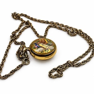 May include: A gold-toned pocket watch on a matching metal chain. The watch face features a colorful enamel inlay depicting a guitar, a lyre, and musical notes against a gold background. The circular case has a winding crown at the top.