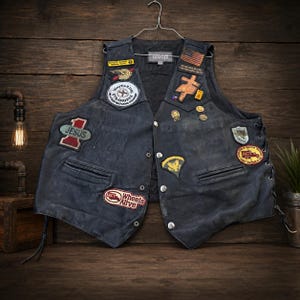 May include: A black leather vest featuring various embroidered patches and metal pins. The patches include text such as Jesus, Wheels Alive, and Wayfaring Strangers. The vest has silver-tone metal snap buttons, side lacing, and two front pockets.