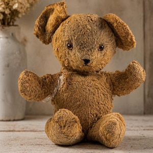 Puede incluir: Un oso de peluche marrón con pelaje texturizado y desgastado, ojos de color marrón oscuro y nariz cosida. El oso tiene una oreja doblada hacia abajo, un parche de reparación visible en el hombro derecho y está sentado con los brazos extendidos.
