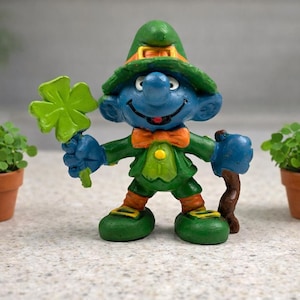 Op de afbeelding: Blauw beeldje van een kabouter met een groene klaver en een houten staf. De kabouter draagt een groene hoed, jas en schoenen, met een oranje strik. Twee kleine potplanten flankeren het beeldje.