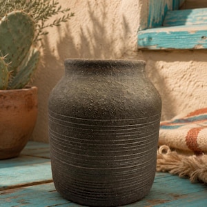 May include: A dark charcoal gray ceramic vase with a rough, textured surface and a series of horizontal incised lines encircling the lower two-thirds of the body. The vase has a wide, rounded shoulder and a short, straight neck with an open top.