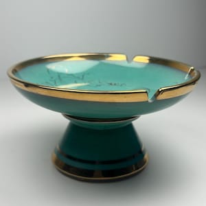以下が含まれることがあります： 金色の縁とベースのアクセントが付いた、ターコイズ色のセラミック灰皿。灰皿は、タバコを置くための小さな切り欠きが付いた円形のボウル型をしています。ベースは、金色のバンドが付いた台座のデザインです。