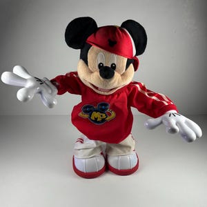 May include: A plush Mickey Mouse toy wearing a red baseball cap and a red and white athletic-style outfit. The toy has large white gloves and shoes with red accents. The outfit features the letters "MB" in a logo on the chest.