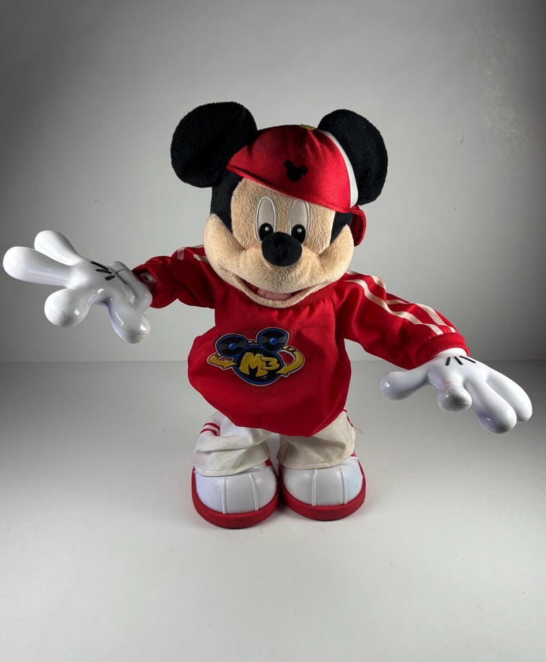 May include: A plush Mickey Mouse toy wearing a red baseball cap and a red and white athletic-style outfit. The toy has large white gloves and shoes with red accents. The outfit features the letters "MB" in a logo on the chest.
