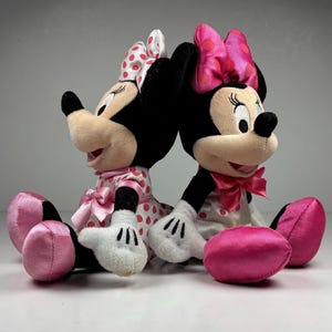 Op de afbeelding: Twee Minnie Mouse pluche speelgoed, rug aan rug. Elk pluche heeft zwarte oren, een roze strik en een roze en witte polkadot jurk. Ze hebben witte handschoenen en roze schoenen. De achtergrond is een effen wit oppervlak.