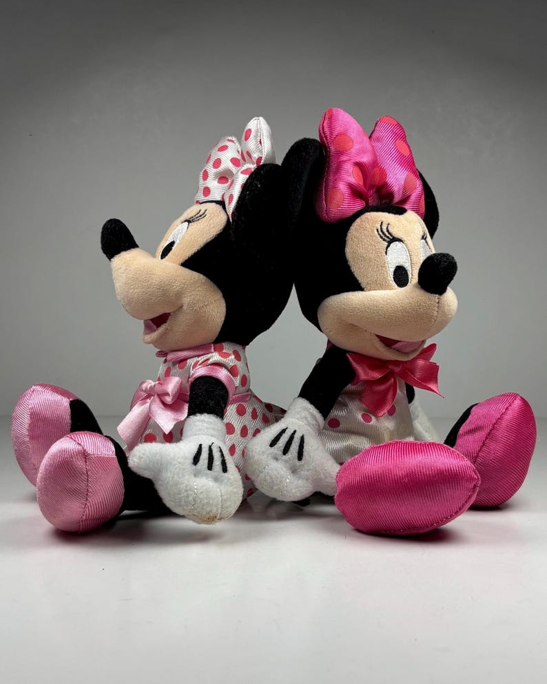 Op de afbeelding: Twee Minnie Mouse pluche speelgoed, rug aan rug. Elk pluche heeft zwarte oren, een roze strik en een roze en witte polkadot jurk. Ze hebben witte handschoenen en roze schoenen. De achtergrond is een effen wit oppervlak.