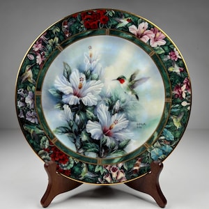 Può includere: Piatto decorativo con un dipinto dettagliato di fiori di ibisco bianchi e un colibrì. Il piatto ha un bordo verde con accenti floreali e colibrì e un bordo dorato. La firma dell'artista, "LENA LIN", è visibile.