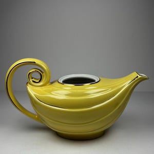 May include: A yellow ceramic teapot with a curved handle and spout. The teapot has a gold trim around the handle and spout, and a white rim around the opening. The teapot has a unique, curved shape.