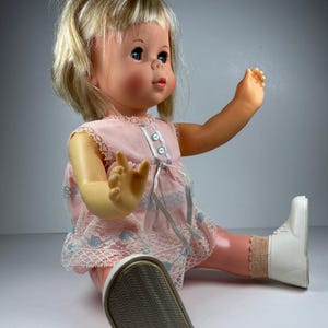 Puede incluir: Una muñeca vintage con cabello rubio, ojos azules y un vestido rosa con ribete de encaje. La muñeca lleva botas blancas y está sentada con un brazo levantado. El vestido de la muñeca tiene detalles florales azules.