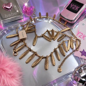 May include: A gold-tone metal necklace featuring a series of textured, leaf-shaped pendants hanging from a delicate chain. The necklace includes a small rectangular tag labeled Talbots and a circular charm with the letter T at the end of the extender.