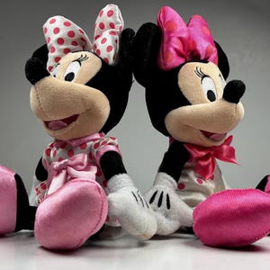 Op de afbeelding: Twee Minnie Mouse pluche poppen. Elke pop heeft zwarte oren, een bruin gezicht en een grote strik met rode stippen. Ze dragen roze jurken met witte stippen, roze strikken en roze schoenen. De poppen zitten naast elkaar.