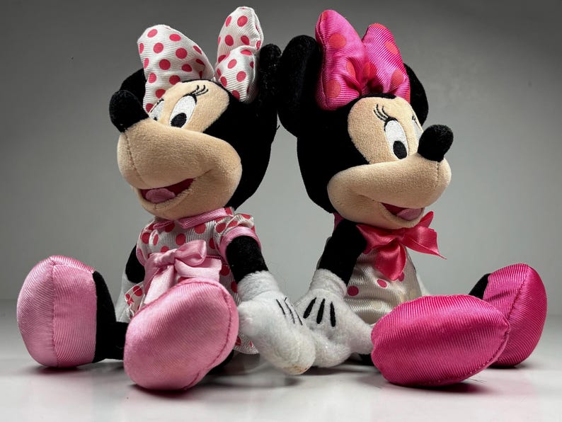 Op de afbeelding: Twee Minnie Mouse pluche poppen. Elke pop heeft zwarte oren, een bruin gezicht en een grote strik met rode stippen. Ze dragen roze jurken met witte stippen, roze strikken en roze schoenen. De poppen zitten naast elkaar.