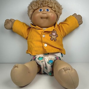 Può includere: Bambola vintage Cabbage Patch Kid con pelle abbronzata, occhi blu e acconciatura riccia castano chiaro. La bambola indossa una giacca di velluto a coste gialla con una toppa a forma di orsetto e un pannolino con motivi.