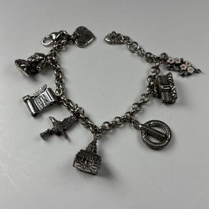 Pu&ograve; includere: Un braccialetto con ciondoli in argento con vari ciondoli, tra cui un cuore, un busto, una pergamena, un uccello, un edificio, un fiore, un pianoforte e un banjo. Il braccialetto &egrave; su una superficie bianca.