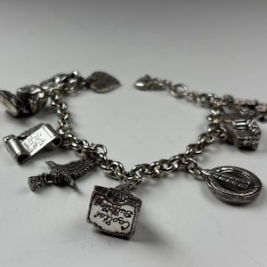 Pu&ograve; includere: Bracciale con ciondoli in argento con pi&ugrave; ciondoli dettagliati. I ciondoli includono una pergamena, un'aquila, un cartello di un edificio con la scritta "Capitol Building" e altri elementi decorativi. Il bracciale &egrave; su una superficie bianca.