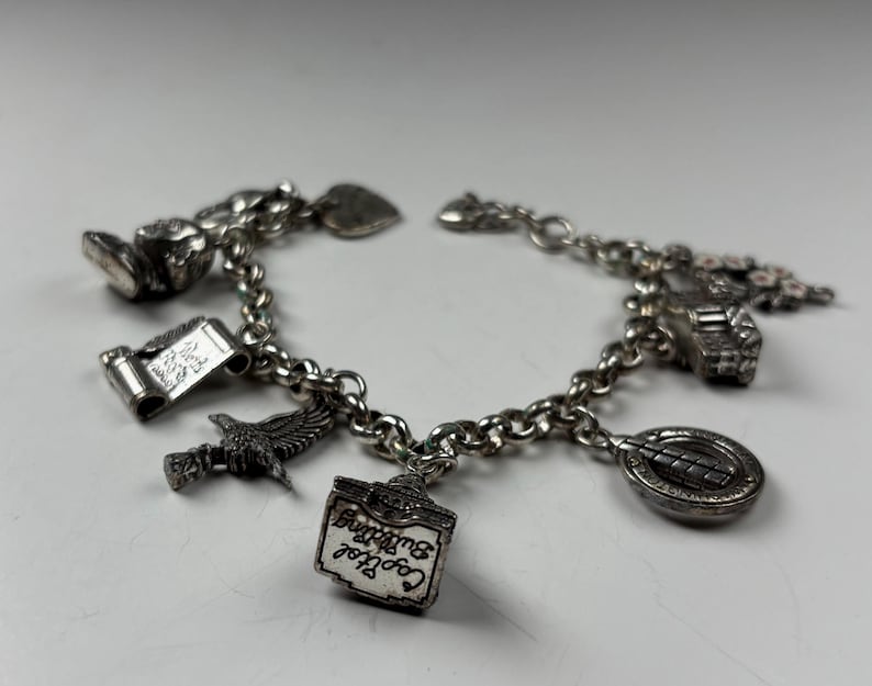 Pu&ograve; includere: Bracciale con ciondoli in argento con pi&ugrave; ciondoli dettagliati. I ciondoli includono una pergamena, un'aquila, un cartello di un edificio con la scritta "Capitol Building" e altri elementi decorativi. Il bracciale &egrave; su una superficie bianca.
