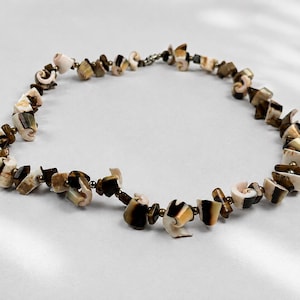 Puede incluir: Un collar que presenta una serie de fragmentos de concha irregulares y pulidos en tonos crema, bronceado y marrón oscuro. Las piezas de concha están intercaladas con pequeñas cuentas redondas de tono metálico, creando un hilo circular texturizado.
