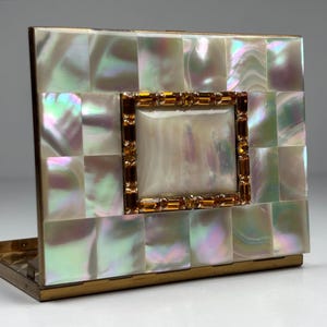May include: A vintage compact mirror with a mother-of-pearl mosaic design. The square compact features a central rectangular frame adorned with amber-colored rhinestones. The edges are trimmed with gold-toned metal.