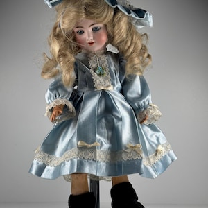 May include: A porcelain doll with blonde hair wearing a light blue satin dress with cream lace trim, small bows, and a matching bonnet. The doll features blue eyes, painted facial details, and black shoes with ribbons tied around the ankles.