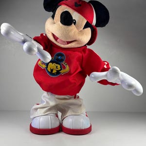 May include: A Mickey Mouse plush toy wearing a red and white outfit, including a red cap and sneakers. The red shirt has a logo with the letters "M3". The toy has white arms and is posed with arms outstretched.